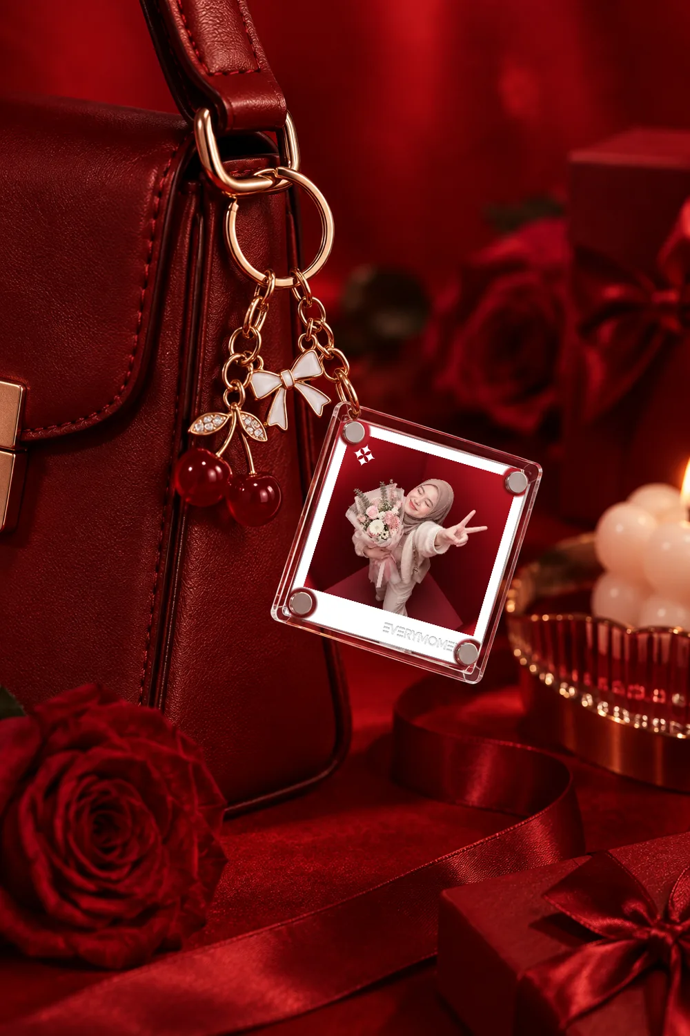 A clear acrylic photo keyring hanging from a burgundy leather handbag on a gold ring, beside a white bow charm and a cherry charm. The photograph inside shows a guest holding a bouquet. Red roses and a candle sit behind.
