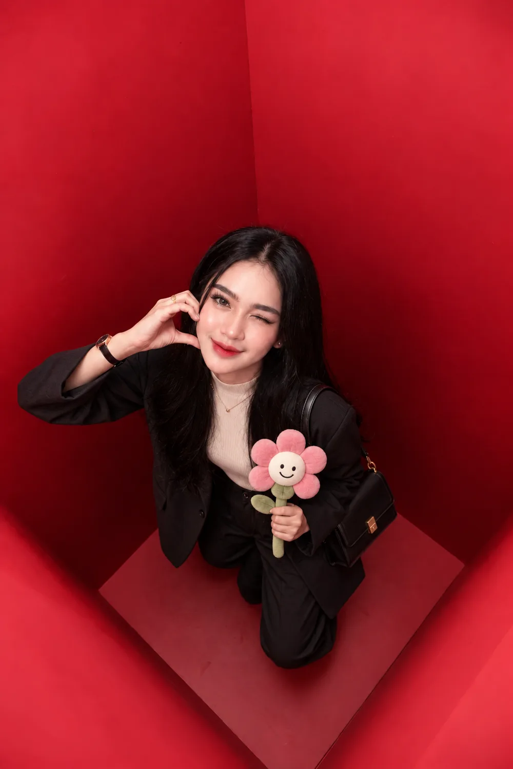 A guest in a black blazer photographed from directly above inside the red booth, holding a small smiling flower toy and touching her face.