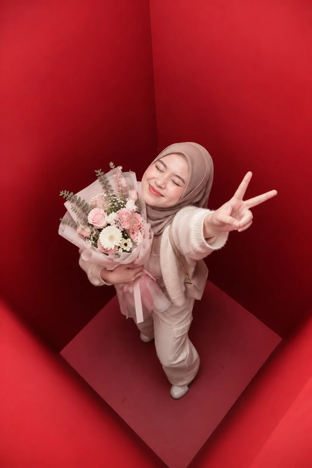 A guest in a hijab photographed from directly above inside the red booth, holding a bouquet and raising a peace sign to the camera.
