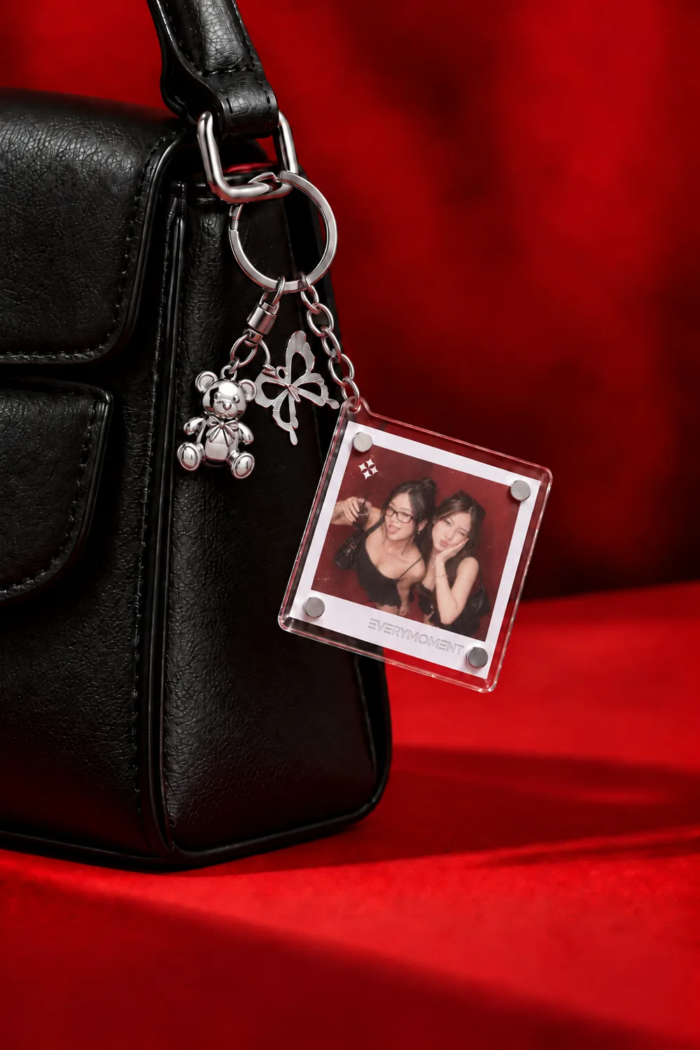 A clear acrylic photo keyring hanging from a black leather handbag beside a silver bow and teddy bear charm, on red velvet. The photograph inside shows two guests posing cheek to cheek.