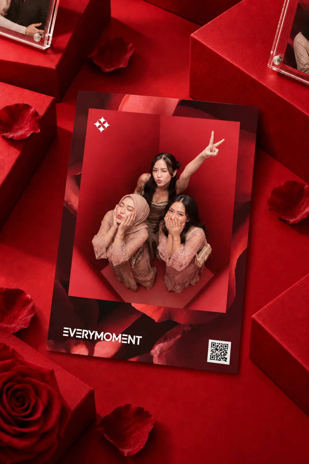 A printed photograph propped among red gift boxes and rose petals. It shows three guests in pink lace, one wearing a hijab, posing inside the red booth, and carries the EVERYMOMENT wordmark and a QR code.