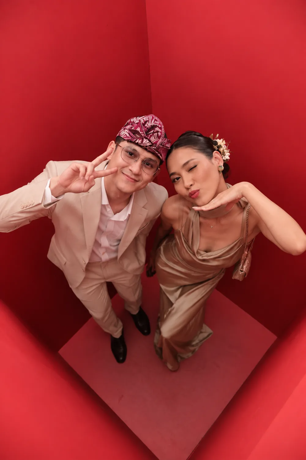 Two guests in formal dress photographed from directly above inside the red booth, one in a patterned cap and cream suit, the other in a gold gown, both raising peace signs.
