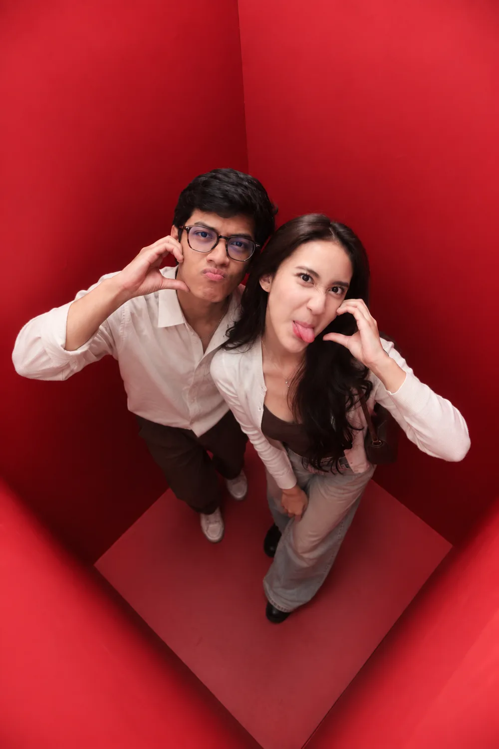 Two guests in cream formal dress photographed from directly above inside the red booth, each framing their face with a raised hand.